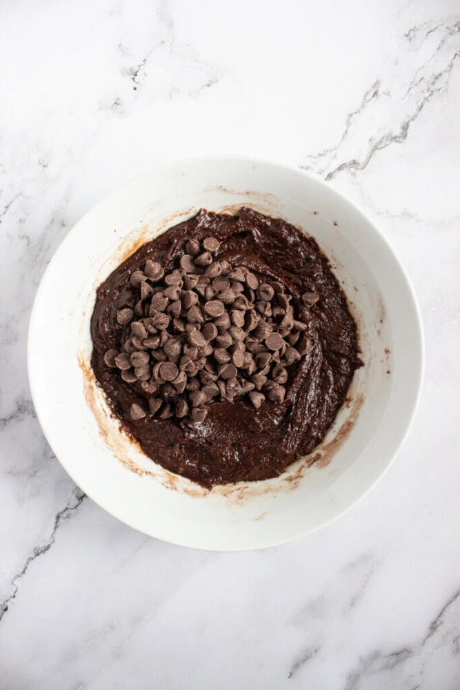 A white bowl filled with chocolate brownie batter topped with a pile of chocolate chips, placed on a marble surface.