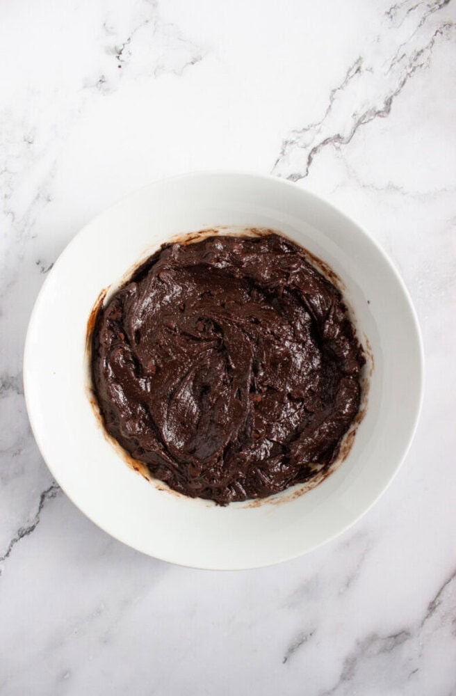 A white bowl filled with thick, dark chocolate batter sits on a marble surface.