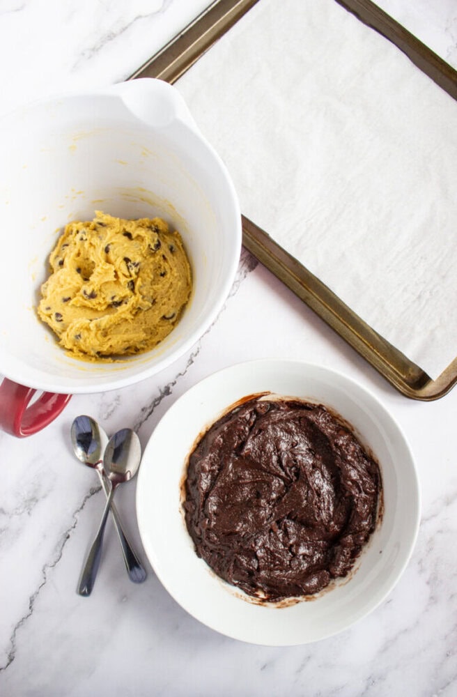 Two bowls of cookie dough, one chocolate chip and one brownie, sit beside two spoons and a parchment-lined baking sheet on a marble counter.
