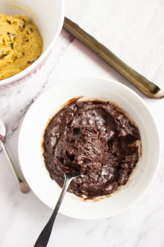 A spoon holding chocolate batter over a white bowl sits next to another bowl with yellow dough, on a white marble surface.