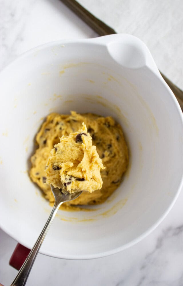 A spoon holding chocolate chip cookie dough over a white mixing bowl, with more dough remaining in the bowl on a marble countertop.