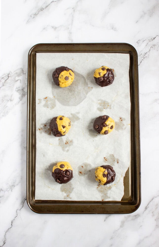 Six unbaked chocolate and vanilla swirl cookie dough balls with chocolate chips on a parchment-lined baking tray, set on a white marble surface.