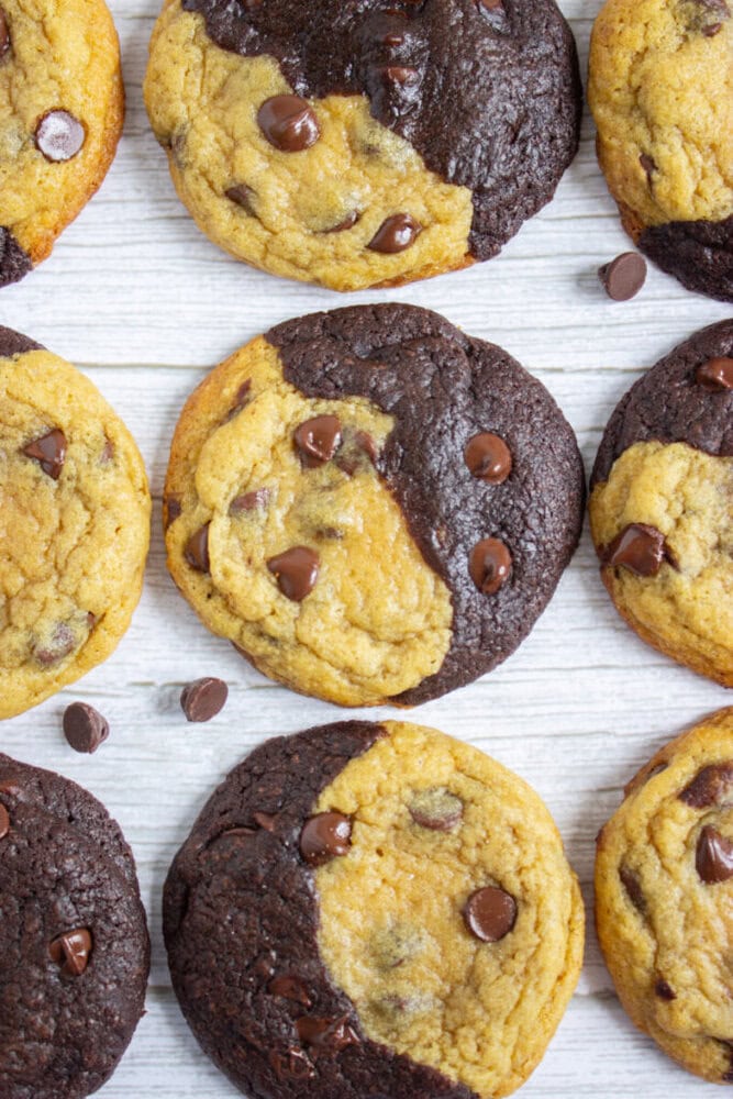 Chocolate chip cookies with a half-vanilla, half-chocolate dough pattern arranged on a white wooden surface, with chocolate chips scattered nearby.