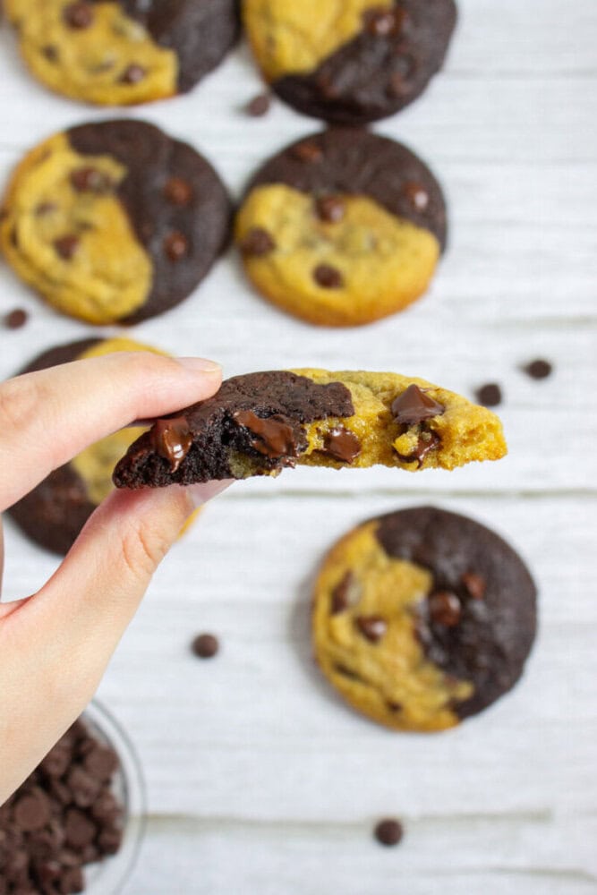 A hand holds a half-eaten chocolate chip cookie that is half chocolate and half regular dough, with whole cookies and chocolate chips in the background.