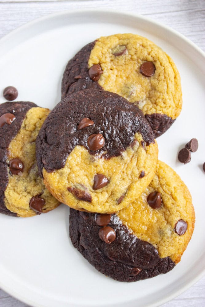 Four large half-chocolate, half-vanilla chocolate chip cookies are arranged on a white plate, with a few loose chocolate chips nearby.