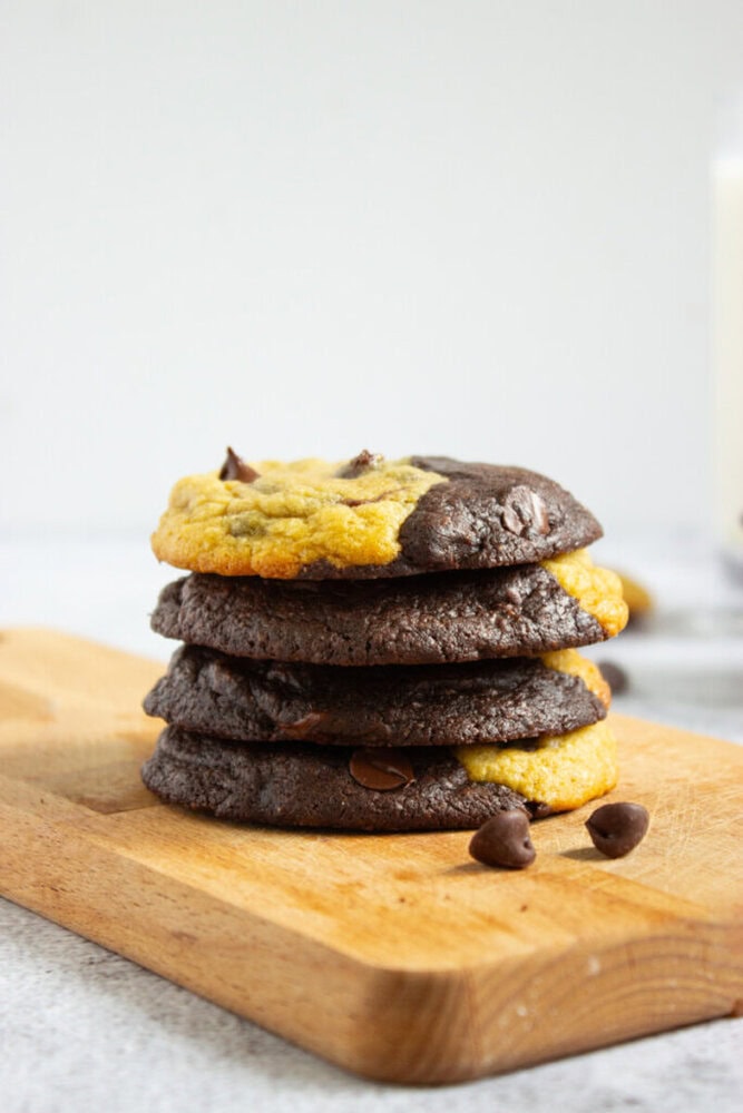 A stack of four black and yellow half-and-half cookies with chocolate chips on a wooden cutting board, with a few chocolate chips scattered nearby.
