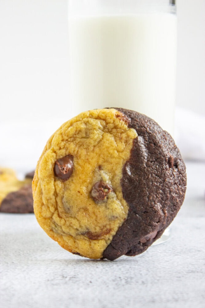 A half-chocolate, half-vanilla chip cookie is leaning against a glass of milk, with another similar cookie blurred in the background.