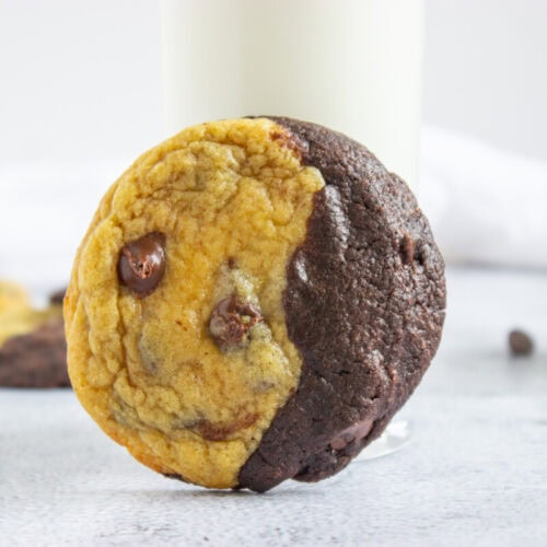 A half-chocolate, half-chocolate chip cookie is propped against a glass of milk, with more cookies and chocolate chips in the background.