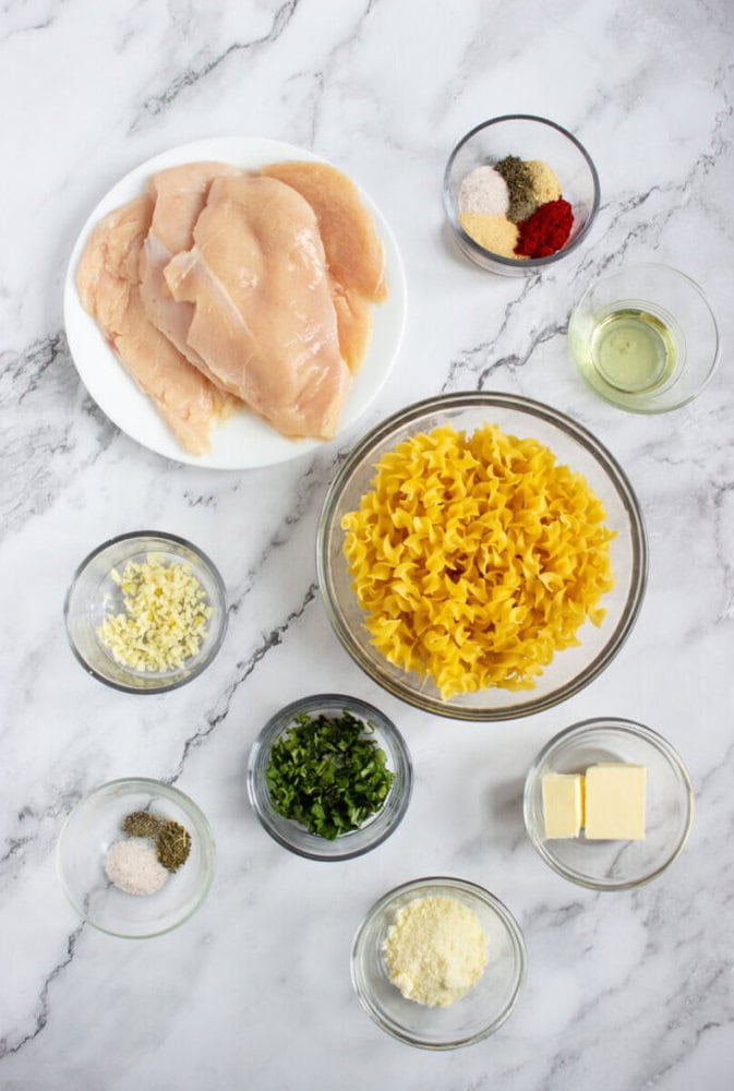 Ingredients arranged on a marble surface include raw chicken breasts, egg noodles, chopped garlic, herbs, spices, oil, butter, and grated cheese in separate bowls.