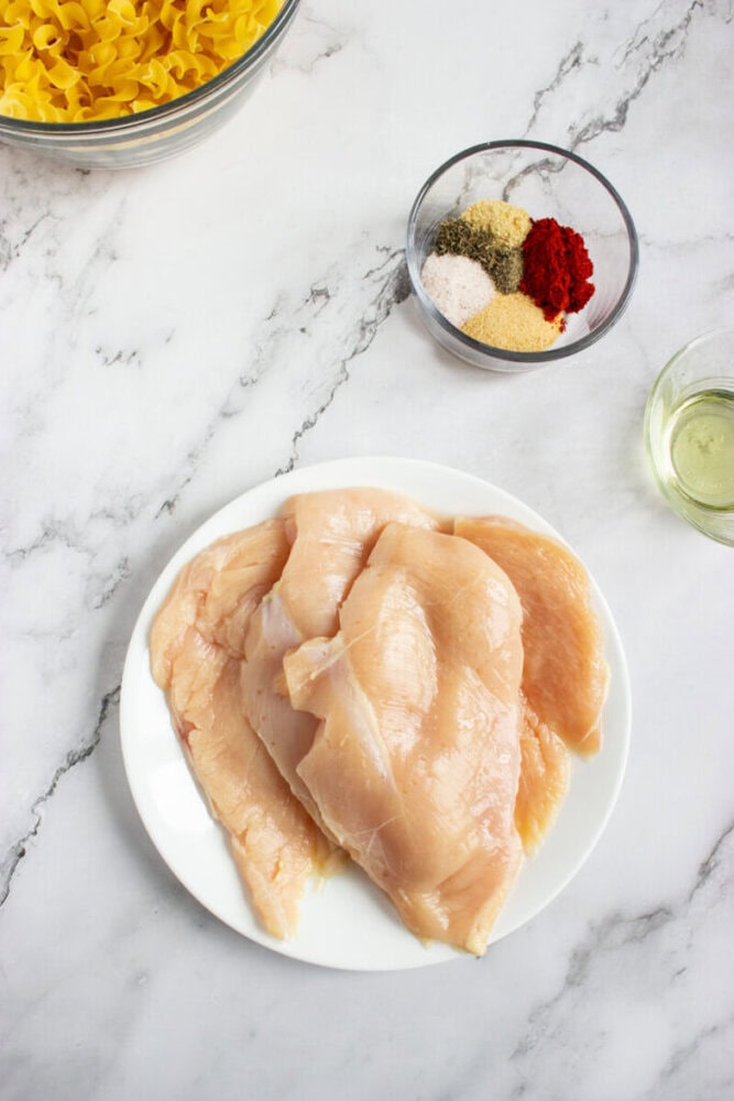 Raw chicken breasts on a white plate, a bowl of pasta, a bowl of mixed spices, and a small glass of oil arranged on a marble countertop.