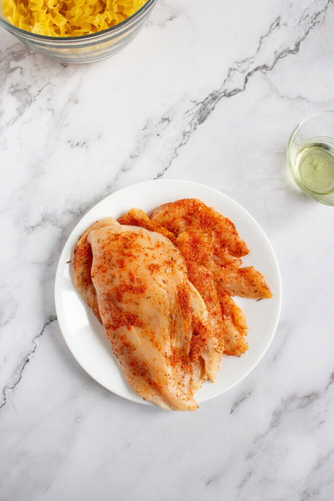 Raw chicken breasts seasoned with spices are arranged on a white plate, with a bowl of uncooked pasta and a small dish of oil nearby on a marble surface.