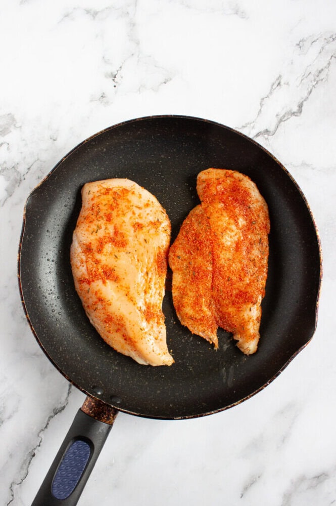 Two raw, seasoned chicken breasts in a black frying pan on a white marble surface.