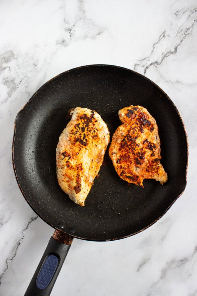 Two seasoned chicken breasts cooking in a black frying pan on a marble countertop.