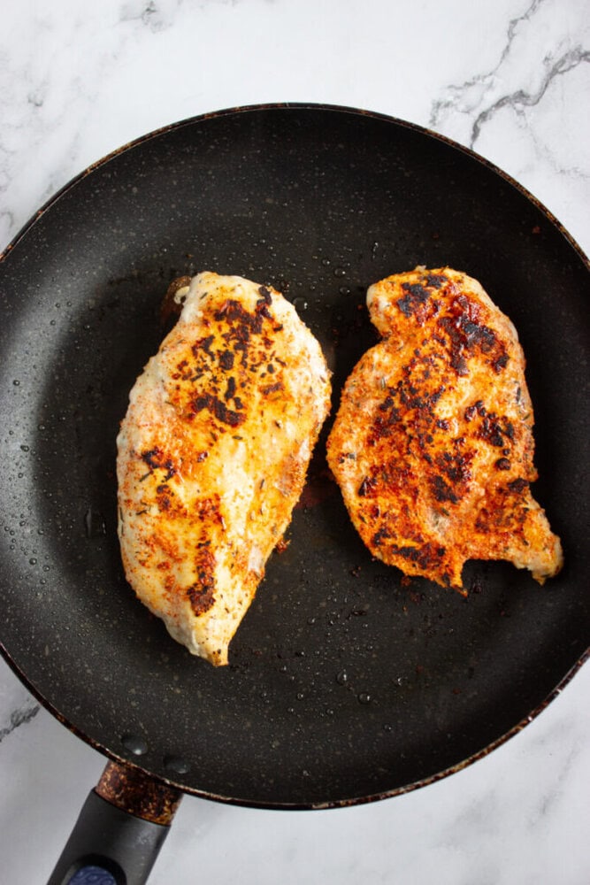 Two cooked, seasoned chicken breasts in a black frying pan on a marble surface.