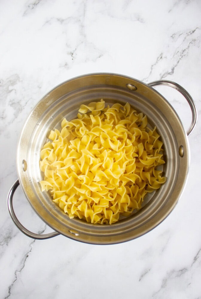 A stainless steel colander filled with cooked egg noodles sits on a white marble surface.