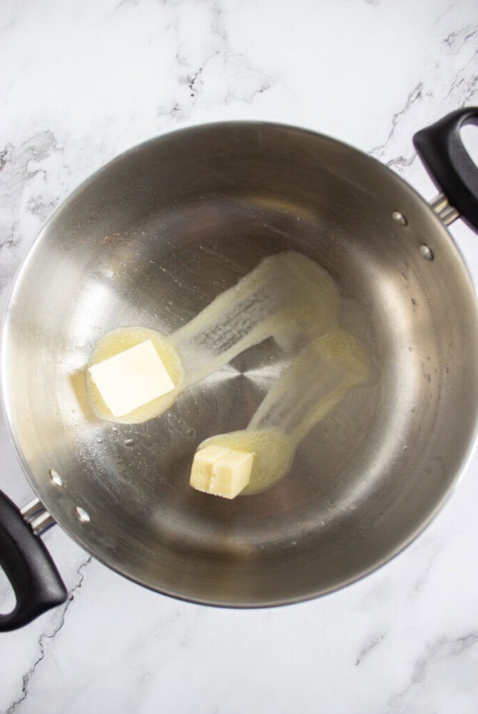 A stainless steel pot with two sticks of butter partially melted on the bottom, placed on a white marble surface.