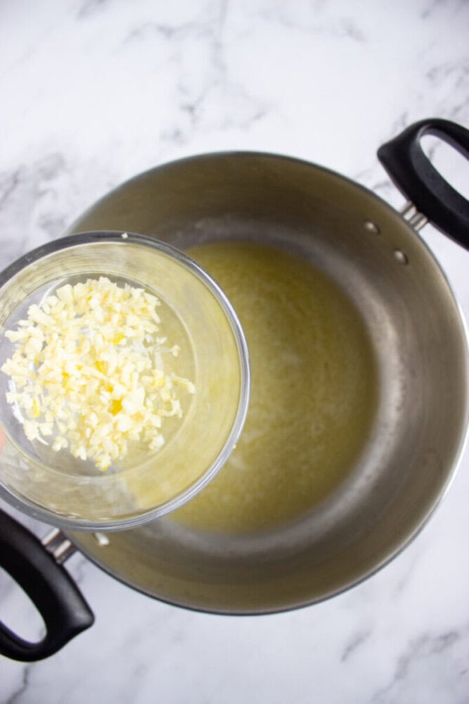 A glass bowl of chopped garlic is about to be added to a large pot with melted butter on a marble countertop.