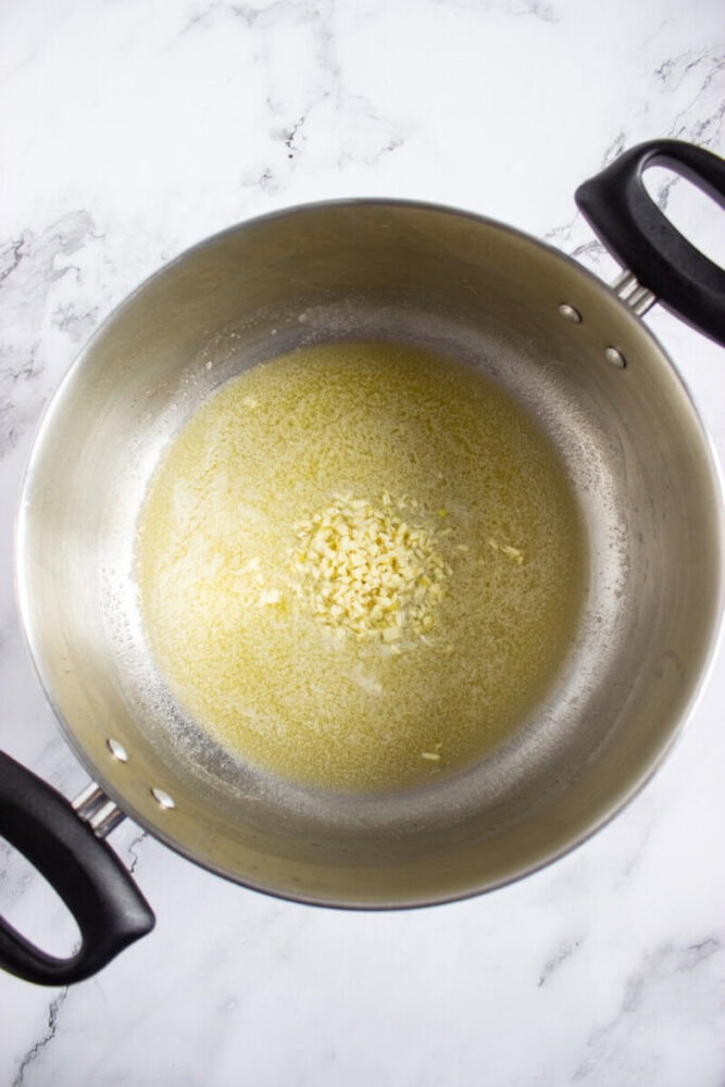 Minced garlic cooking in melted butter in a large stainless steel pot on a white marble countertop.