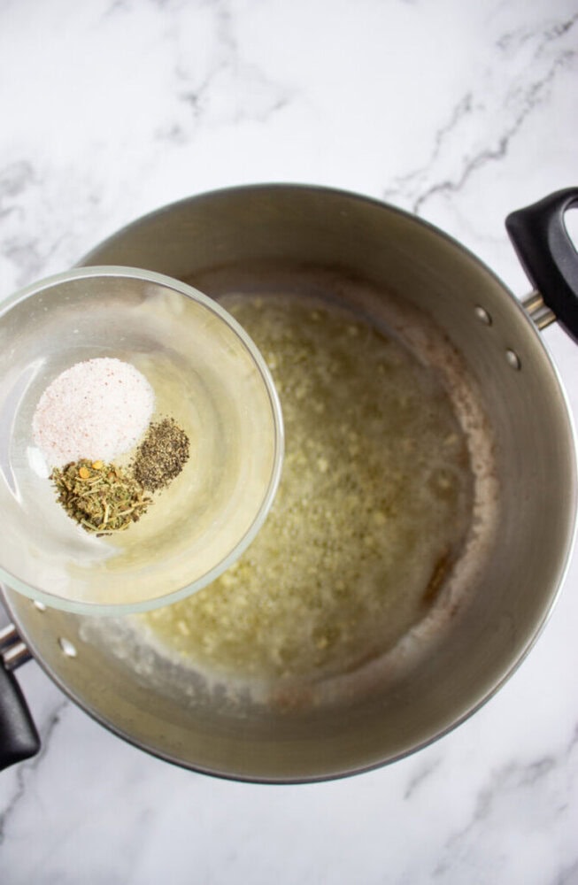 A glass bowl with salt and dried herbs is held above a pot of melted butter on a marble countertop.