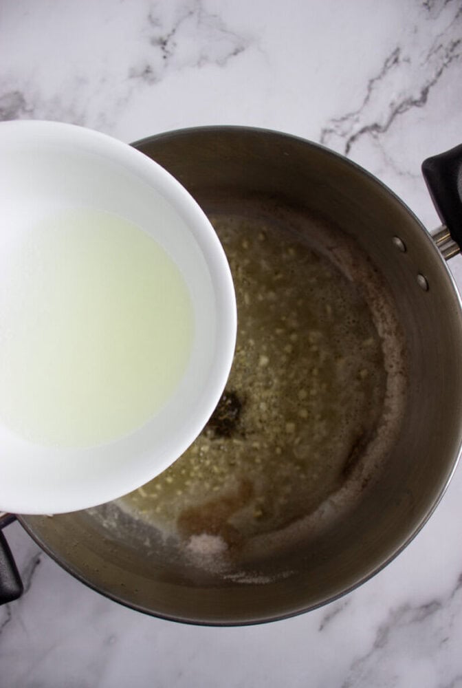 A white bowl with liquid is being poured into a pot containing browned butter on a marble countertop.