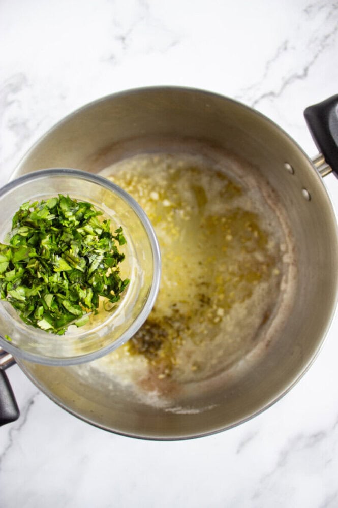 Chopped herbs being added to a pot of melted butter and minced garlic on a marble countertop.