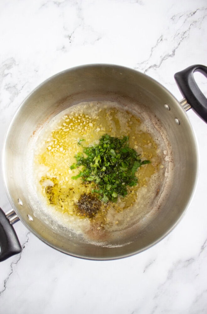 Minced garlic and chopped herbs sautéing in melted butter in a stainless steel pot on a marble countertop.