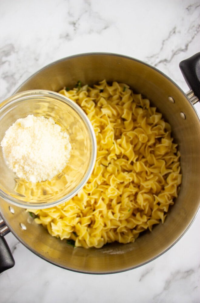 A pot of cooked egg noodles with a small glass bowl of grated cheese being held above the noodles, ready to be added.