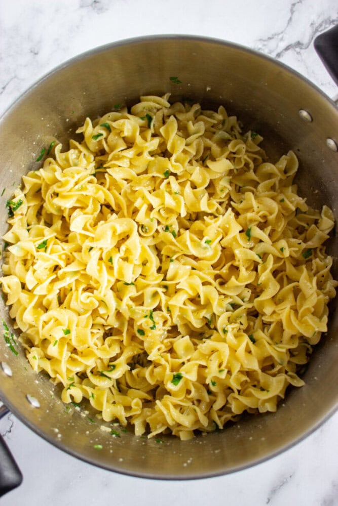 A large pot filled with cooked egg noodles garnished with chopped herbs, sitting on a white marble surface.