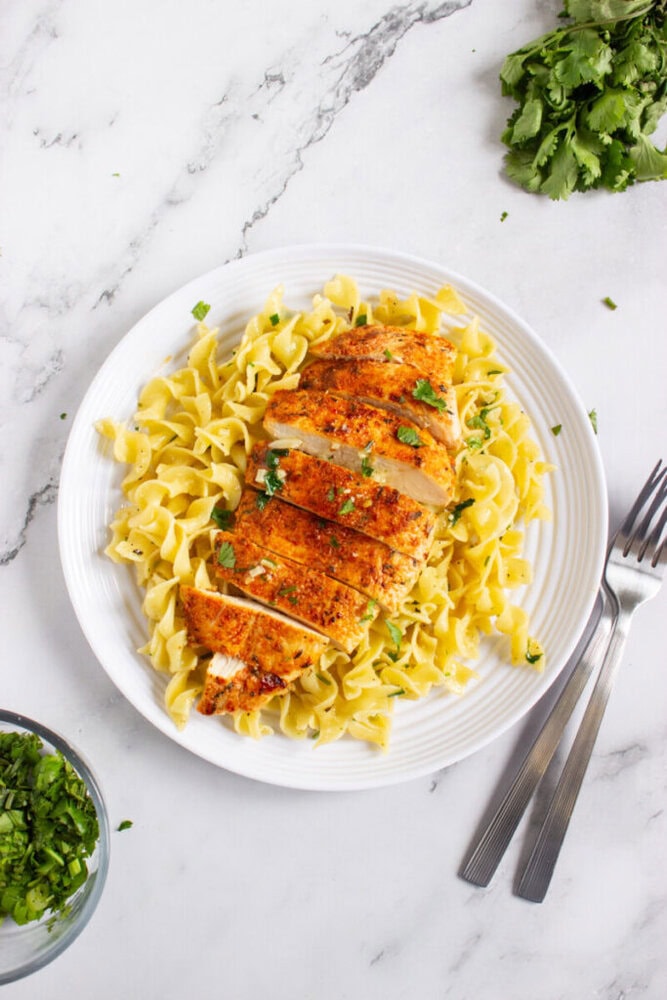A white plate with sliced seasoned chicken breast served over egg noodles, garnished with chopped herbs. Two forks are placed next to the plate on a marble surface.