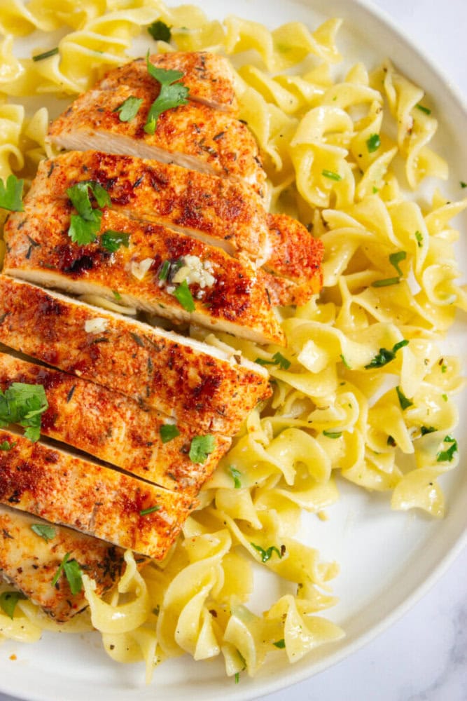 Sliced seasoned chicken breast served on a bed of buttered egg noodles, garnished with chopped parsley on a white plate.