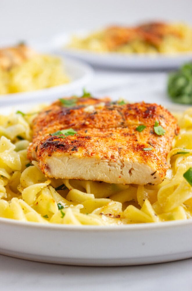A close-up of a seasoned, grilled chicken breast served on top of cooked pasta with a light sauce, garnished with chopped herbs.
