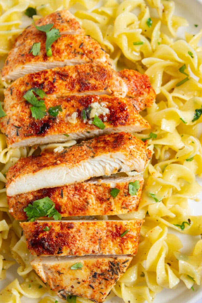 Sliced seasoned chicken breast on a bed of cooked egg noodles, garnished with chopped parsley.