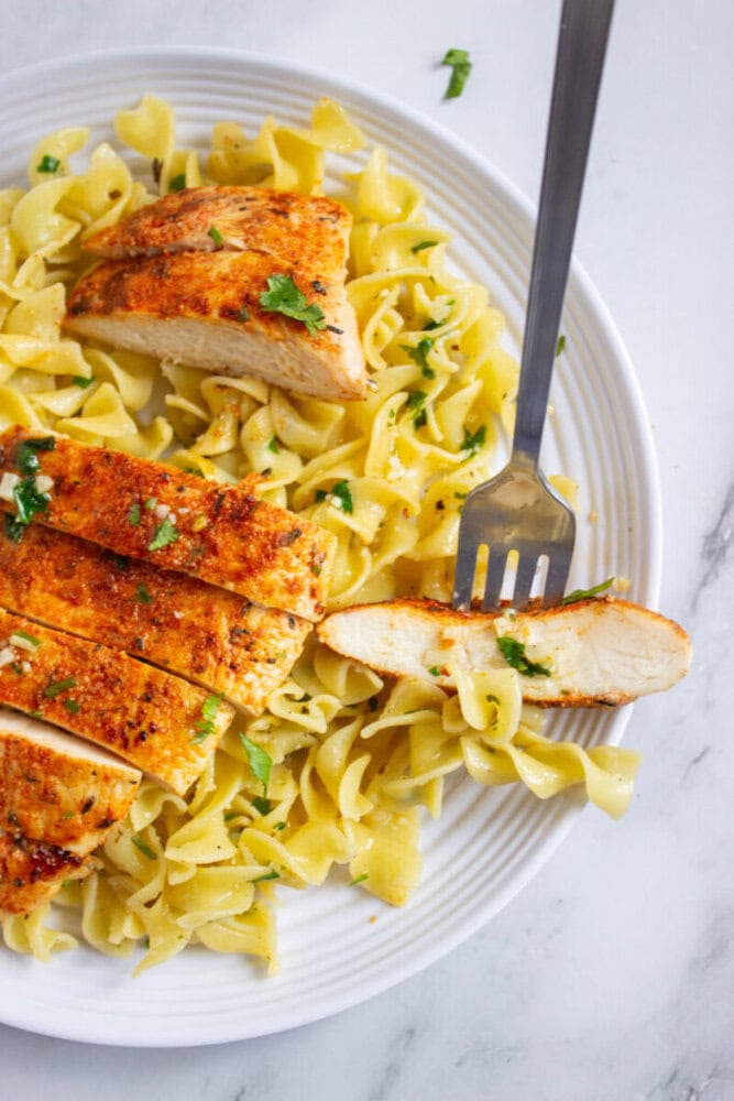 A plate of seasoned, sliced grilled chicken breast served over egg noodles, garnished with chopped herbs, with a fork holding a piece of chicken.