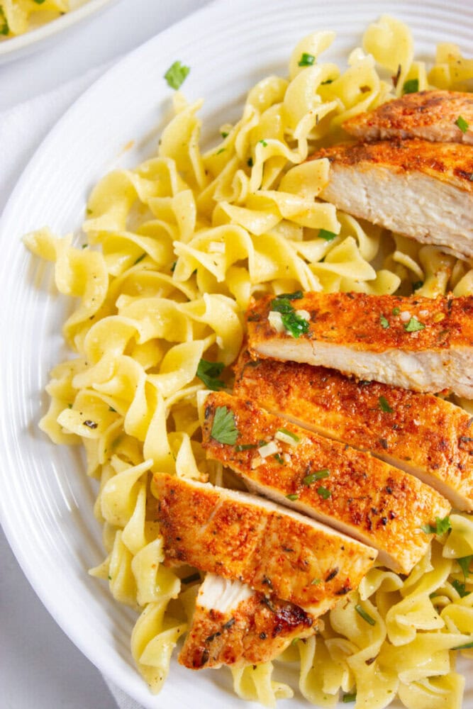 Sliced seasoned chicken breast served over a bed of cooked egg noodles, garnished with chopped herbs on a white plate.