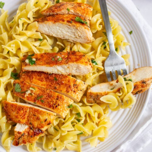 A plate of sliced grilled chicken breast served on top of egg noodles, garnished with chopped herbs, with a fork resting on the plate.