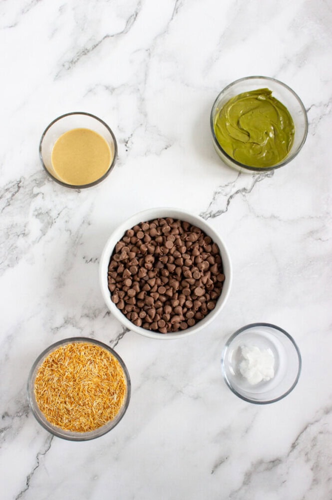 Five bowls on a marble surface containing chocolate chips, green matcha spread, coconut oil, shredded coconut, and a tan nut butter.