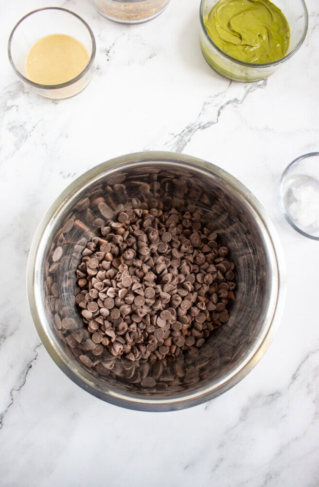 A metal bowl filled with chocolate chips sits on a marble countertop, surrounded by small glass bowls containing different ingredients.