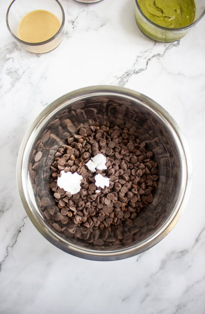 Metal bowl filled with chocolate chips and scoops of coconut oil on a marble countertop, with bowls of green and beige mixtures nearby.