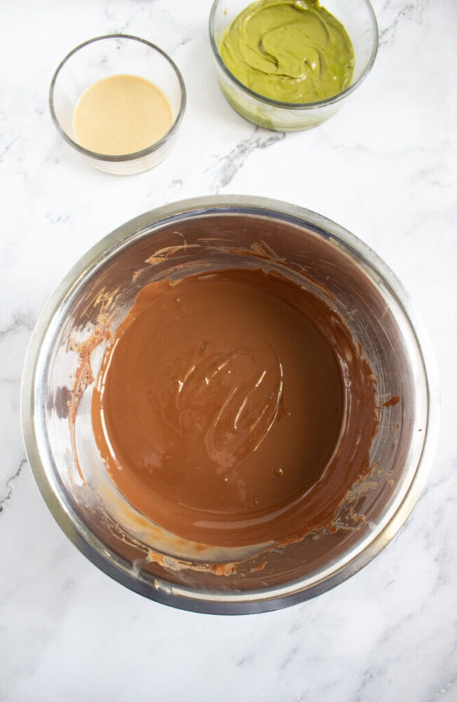 A metal bowl filled with melted chocolate, with two smaller glass bowls containing light beige and green mixtures, all on a white marble surface.