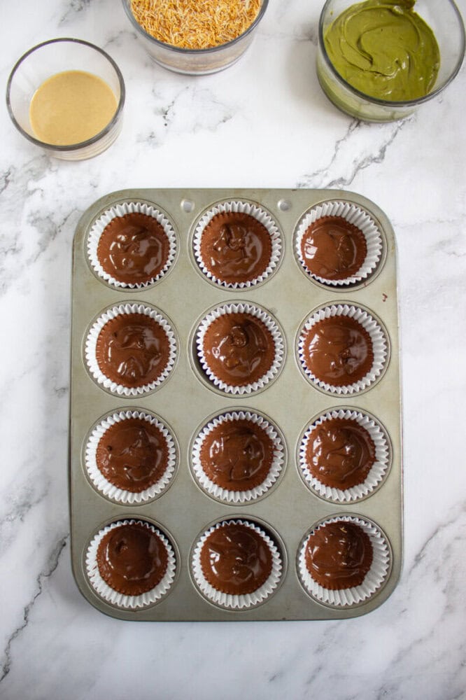 A muffin tin with twelve chocolate batter-filled cupcake liners on a marble surface, with three bowls containing different ingredients nearby.