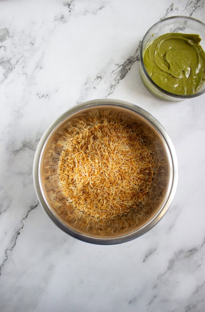 A metal bowl filled with dry, thin vermicelli noodles sits on a marble surface beside a small glass bowl containing green paste.