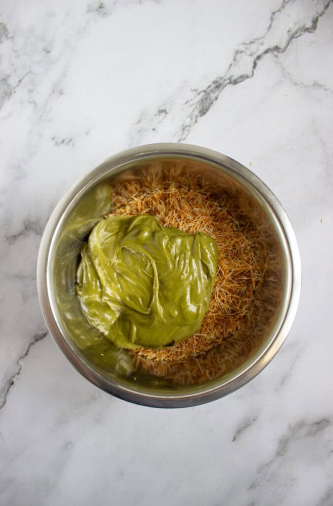 A metal bowl containing a layer of dry vermicelli noodles topped with a thick layer of green paste, placed on a white marble surface.