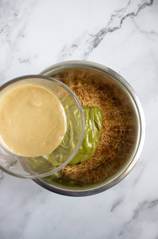 A glass bowl pours a greenish-brown liquid over dry vermicelli noodles in a stainless steel mixing bowl on a white marble surface.