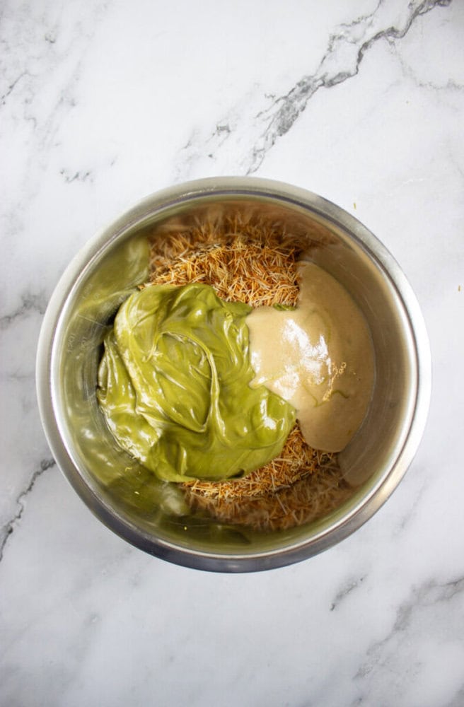 A stainless steel bowl containing dry shredded noodles, green matcha mixture, and light brown tahini, placed on a marble countertop.