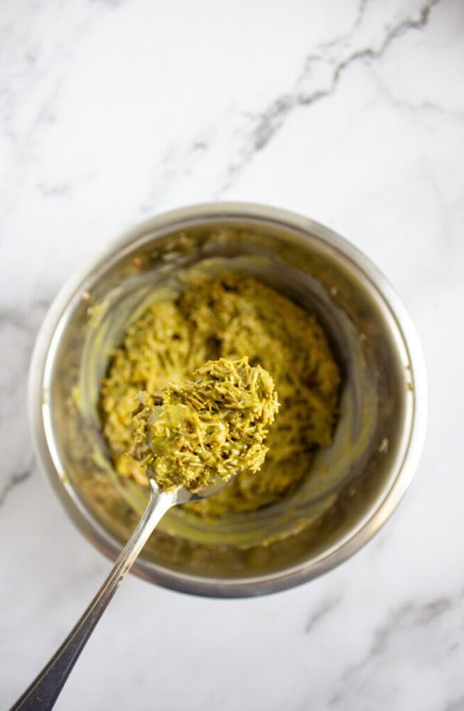 A metal bowl filled with a greenish batter or mixture, with a spoon lifting some of the mixture above the bowl on a marble surface.