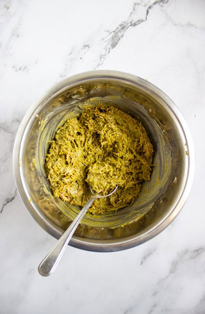 A metal bowl containing a mixture of shredded ingredients with a greenish hue, and a spoon resting inside, on a white marble surface.
