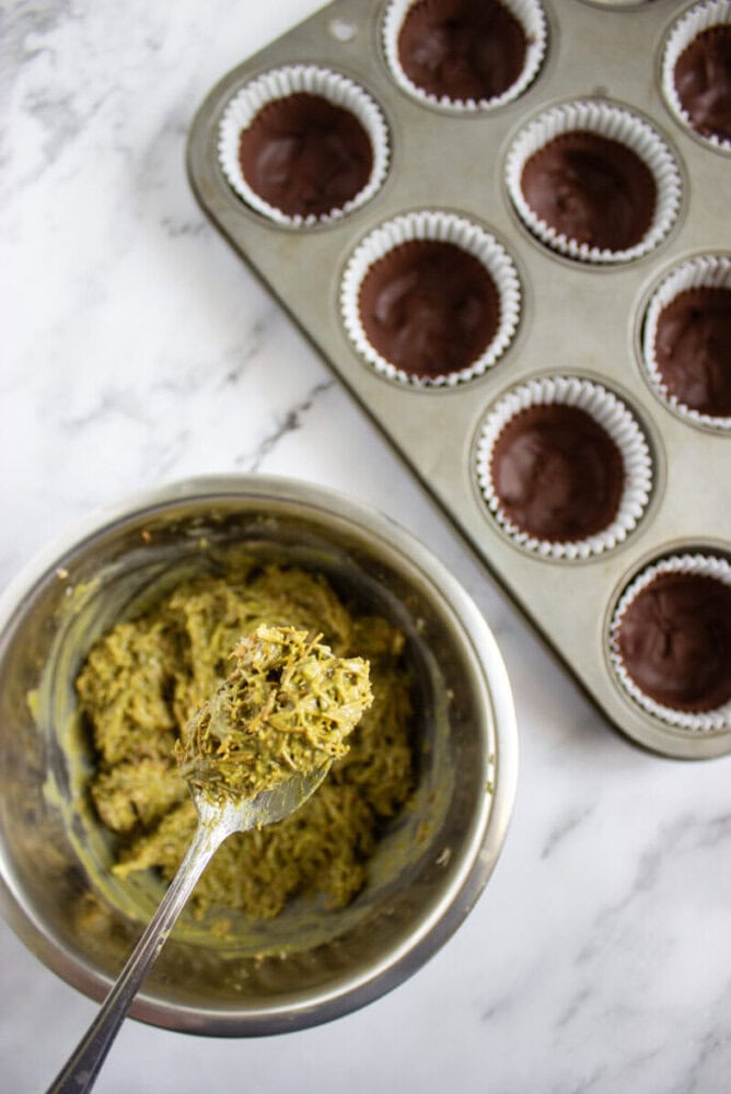 A metal mixing bowl with green cookie dough and a spoon is next to a muffin tin filled with chocolate batter in paper liners on a marble surface.