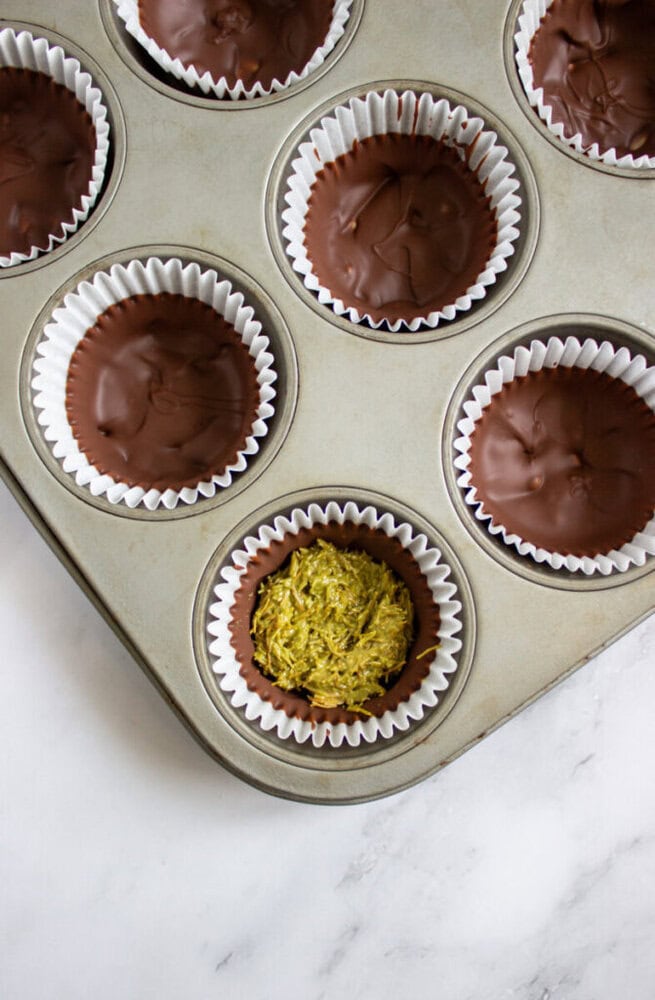A muffin tin with six paper liners, five filled with chocolate-covered treats and one showing a green, textured filling.