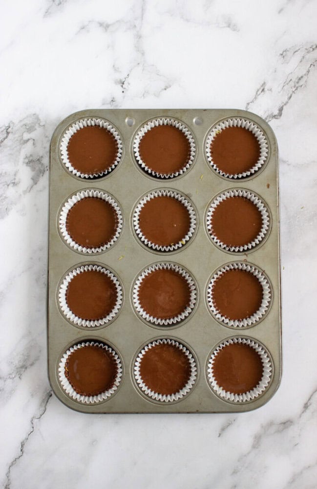 A muffin tin with twelve cups, each filled with chocolate batter in white paper liners, sits on a marble surface.