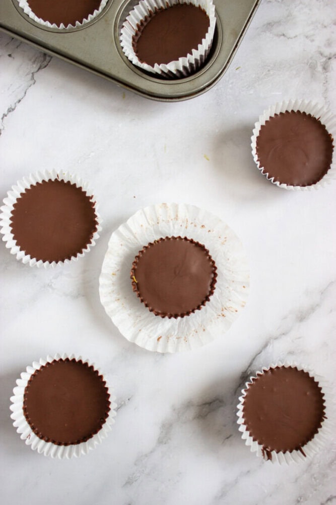 Five chocolate peanut butter cups in white paper liners are arranged on a marble surface, with a muffin tin in the corner holding one cup.
